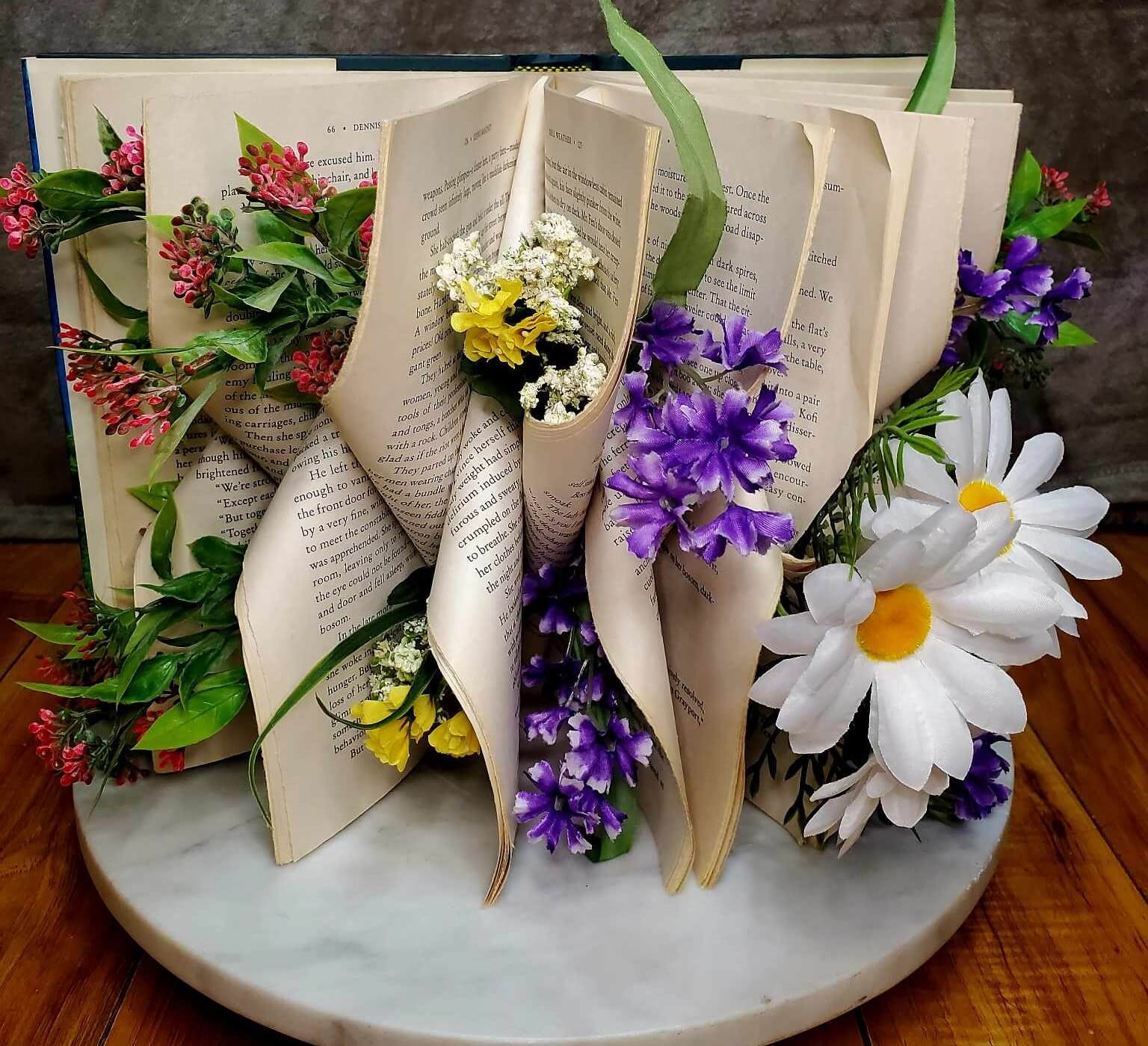 An intricately folded book with small pink, yellow and purple flowers arranged throughout the folds. Two large daisies sit within a fold to the right.