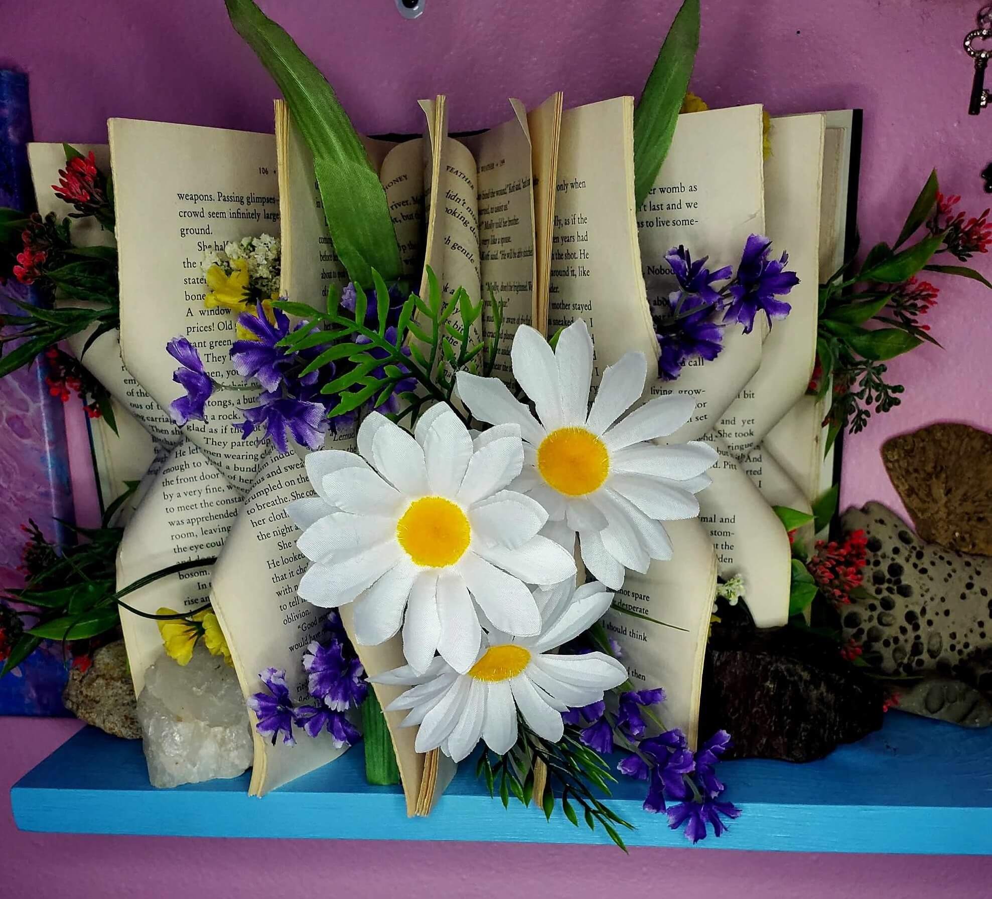 An intricately folded book with three large daisies arranged in the center and small purple flowers and foliage arranged throughout the folds.