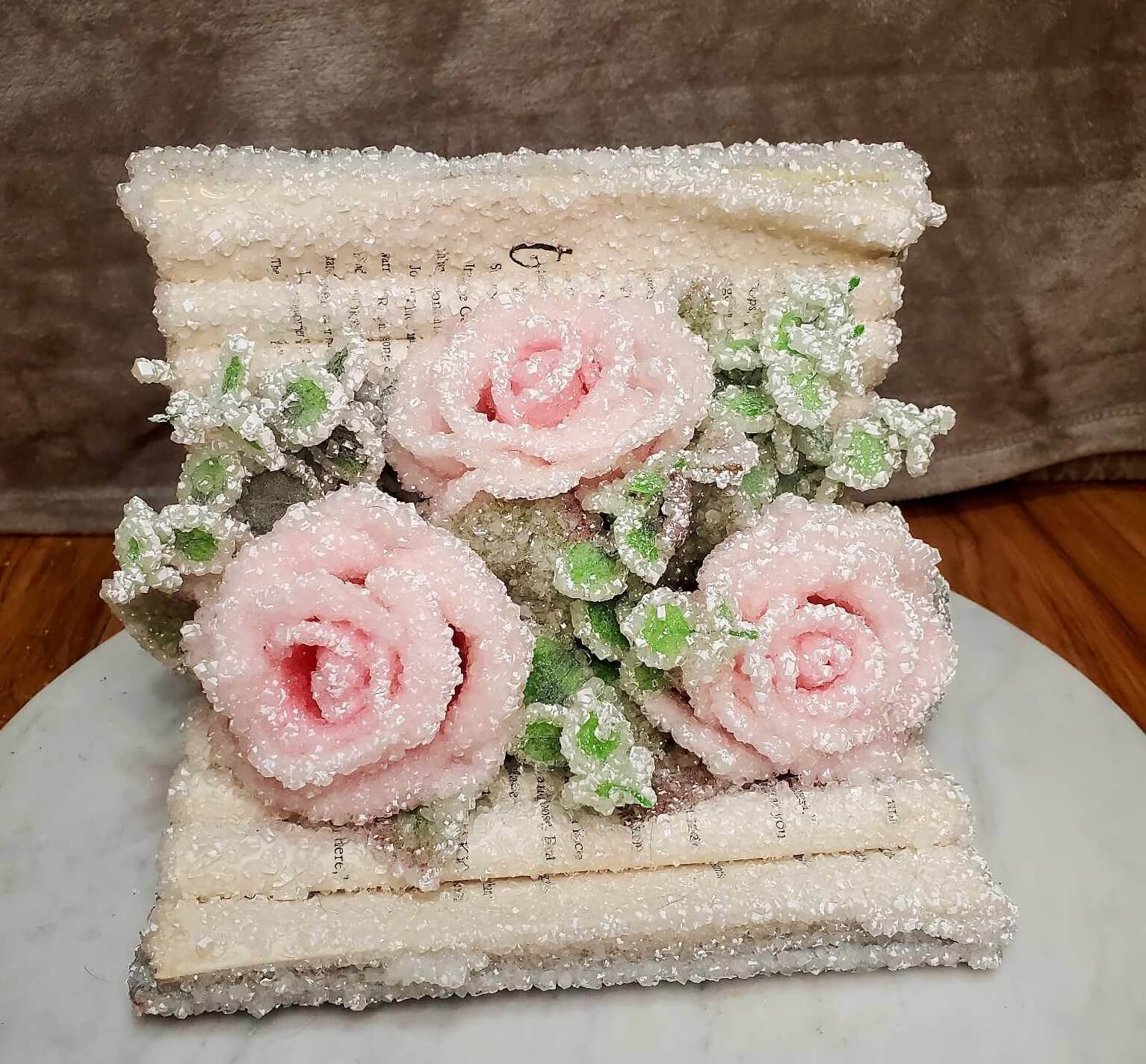 A crystallised folded book opened on its side with three pink roses arranged in the center.