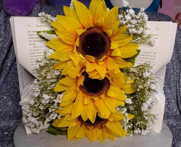 Three large sunflowers appear to grow from the folded page of a book that is standing upright.