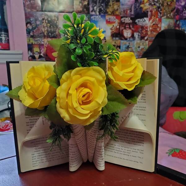 A book with folded pages standing upright with yellow roses emerging from the folds.